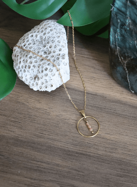 Collier Calista en acier inoxydable doré avec pendentif cercle et perles de pierre de soleil, bijou lumineux de 40 cm posé sur fond beige.