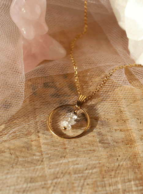 Collier Lucibelle en acier inoxydable doré avec pendentif cercle et cristal de diamant d’Herkimer, bijou raffiné et lumineux présenté sur fond en bois et voile rose poudré.