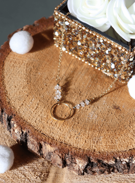 Collier Lucille en acier inoxydable doré avec pendentif cercle et perles de diamant d’Herkimer, bijou lumineux de 40 cm posé sur fond en bois entouré de fleurs blanches.
