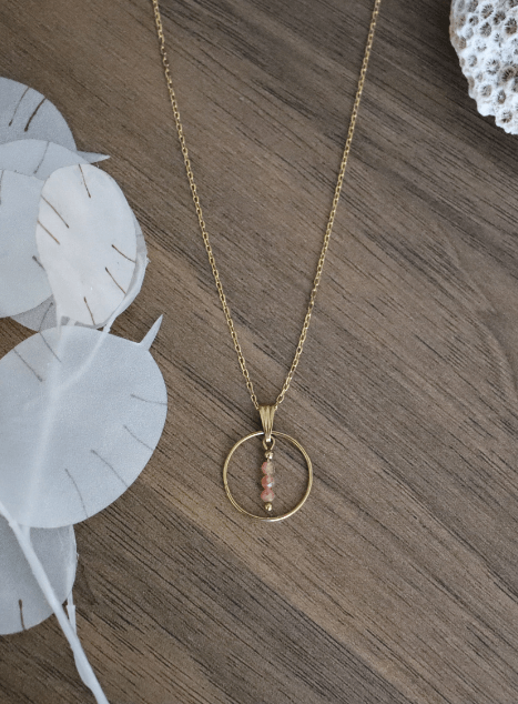 collier-calista-pierre-de-soleil-cercle-acier-inoxydable-dore-40cm.jpg Collier Calista en acier inoxydable doré avec pendentif cercle et perles de pierre de soleil, bijou lumineux de 40 cm posé sur fond beige.