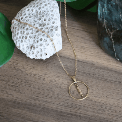 Collier Calista en acier inoxydable doré avec pendentif cercle et perles de pierre de soleil, bijou lumineux de 40 cm posé sur fond beige.