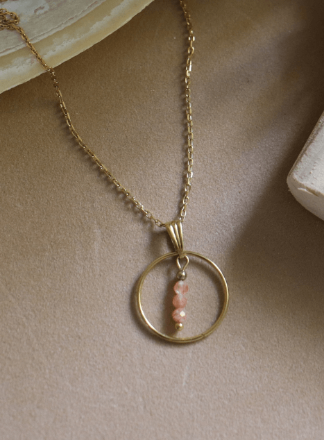 collier-calista-pierre-de-soleil-cercle-acier-inoxydable-dore-40cm.jpg Collier Calista en acier inoxydable doré avec pendentif cercle et perles de pierre de soleil, bijou lumineux de 40 cm posé sur fond beige.