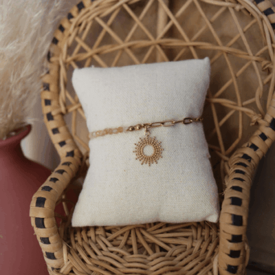 Bracelet Chacha en acier inoxydable doré avec perles de pierre de soleil et pendentif soleil, bijou lumineux présenté sur coussin en lin dans une chaise en rotin.