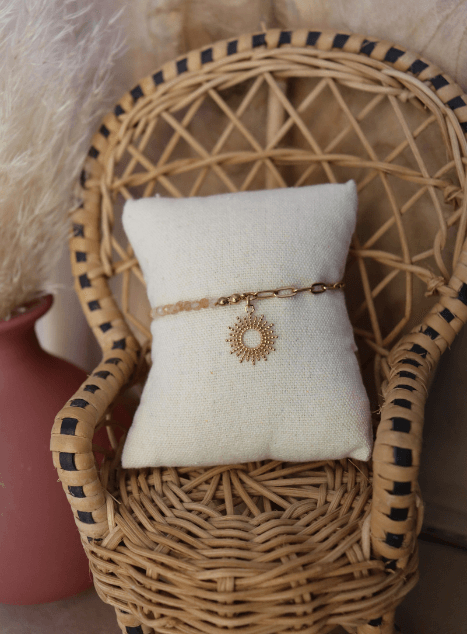 bracelet-chacha-pierre-de-soleil-pendentif-soleil-acier-inoxydable-dore.jpg Bracelet Chacha en acier inoxydable doré avec perles de pierre de soleil et pendentif soleil, bijou lumineux présenté sur coussin en lin dans une chaise en rotin.