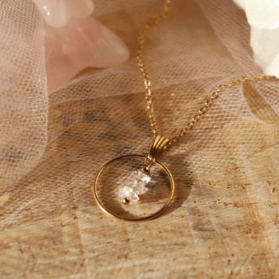 Collier Lucibelle en acier inoxydable doré avec pendentif cercle et cristal de diamant d’Herkimer, bijou raffiné et lumineux présenté sur fond en bois et voile rose poudré.