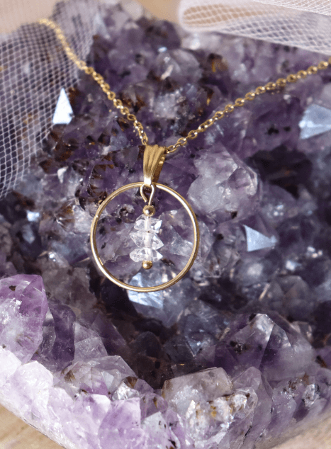 collier-lucibelle-diamant-herkimer-cercle-acier-inoxydable-dore-sur-geode-amethyste.jpg ollier Lucibelle en acier inoxydable doré avec pendentif cercle et cristal de diamant d’Herkimer, posé sur une géode d’améthyste pour un rendu lumineux et raffiné.