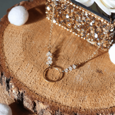 Collier Lucille en acier inoxydable doré avec pendentif cercle et perles de diamant d’Herkimer, bijou lumineux de 40 cm posé sur fond en bois entouré de fleurs blanches.