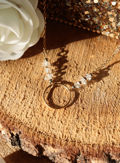 collier-lucille-diamant-herkimer-cercle-acier-inoxydable-dore-40cm.jpg Collier Lucille en acier inoxydable doré avec pendentif cercle et perles de diamant d’Herkimer, bijou lumineux de 40 cm posé sur fond en bois entouré de fleurs blanches.