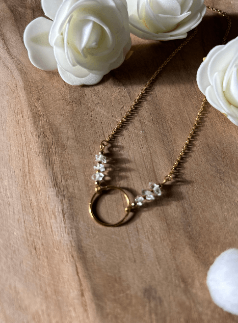 collier-lucille-diamant-herkimer-cercle-acier-inoxydable-dore-40cm.jpg Collier Lucille en acier inoxydable doré avec pendentif cercle et perles de diamant d’Herkimer, bijou lumineux de 40 cm posé sur fond en bois entouré de fleurs blanches.