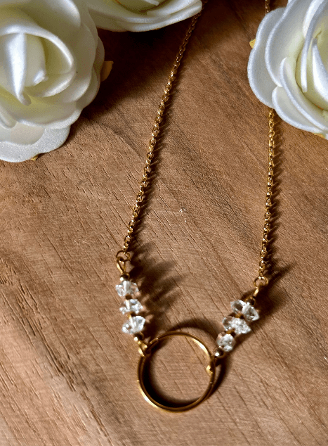 collier-lucille-diamant-herkimer-cercle-acier-inoxydable-dore-40cm.jpg Collier Lucille en acier inoxydable doré avec pendentif cercle et perles de diamant d’Herkimer, bijou lumineux de 40 cm posé sur fond en bois entouré de fleurs blanches.