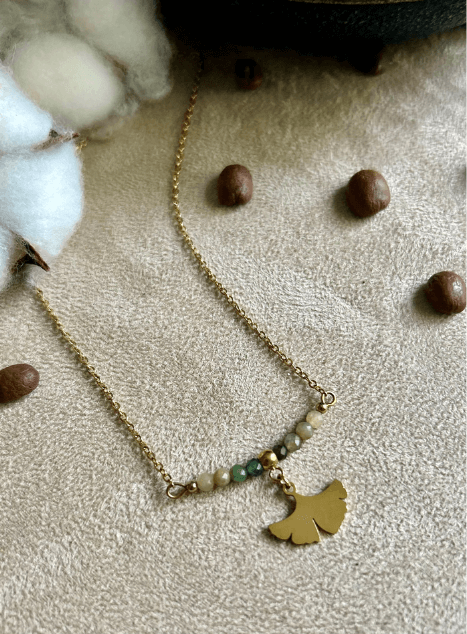Collier Sienna en acier inoxydable doré avec pendentif feuille de ginkgo et perles de jade verte, bijou délicat de 40 cm posé sur fond beige.