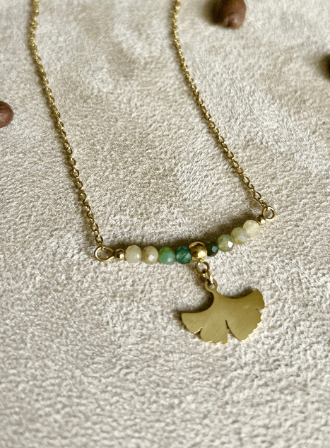 Collier Sienna en acier inoxydable doré avec pendentif feuille de ginkgo et perles de jade verte, bijou délicat de 40 cm posé sur fond beige.