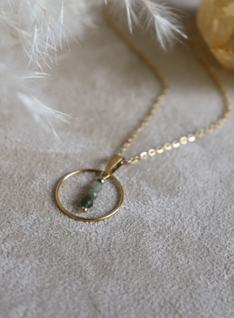 collier-sya-jade-verte-cercle-acier-inoxydable-dore-40cm.jpg Collier Sya en acier inoxydable doré avec pendentif cercle et perles de jade verte, bijou raffiné de 40 cm posé sur fond beige doux.